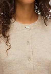 Cheveux bouclés tombant sur les épaules, portant un cardigan en tricot beige boutonné avec quatre boutons clairs, col visible.