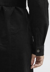 Manteau en velours côtelé noir avec une silhouette ajustée, des manches longues à poignets boutonnés et une taille marquée. Texture remarquable et finition douce visibles.
