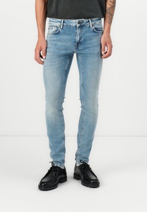 Homme portant un jean slim bleu clair et des chaussures noires à lacets, debout sur un fond blanc.