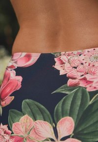 Gėlių rašto maudymosi šortai su tamsiai mėlynu fonu, puikuojasi didelėmis rožinėmis gėlėmis ir žaliomis lapų. Auksinės detalės juosmens srityje.