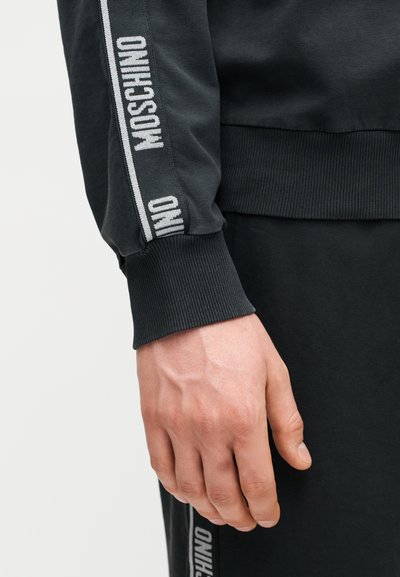 Κοντινό πλάνο ενός χεριού δίπλα σε μαύρο μανίκι και παντελόνι με λευκό κείμενο "MOSCHINO" σε ριγωτό υφασμάτινο τελείωμα.