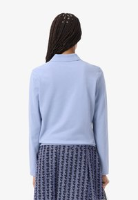 Polo shirt a maniche lunghe azzurro chiaro con colletto pieghevole, texture liscia e vestibilità su misura. Abbinato a una gonna navale a fantasia.