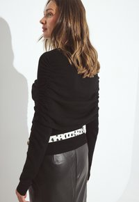 Sieviete ar plecu garuma viļņainiem matiem, tērpusies melnā pieguļošā džemperī, melnā ādas svārkos un baltā jostā ar melniem plankumiem, stāv sānis.