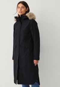 Parka lunga nera con cappuccio bordato in pelliccia, chiusura frontale con zip, due tasche laterali e dettagli con bottoni; realizzata in un materiale resistente alle intemperie.