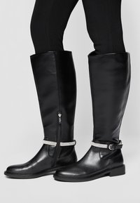 Bottes en cuir noir montantes jusqu'aux genoux avec un design élégant, fermeture éclair et une sangle décorative ornée de strass à la cheville.