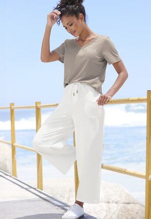Beige v-neck, short-sleeve t-shirt with logo; white, wide-leg trousers with drawstring waist; paired with white sneakers. Ocean and railing background.