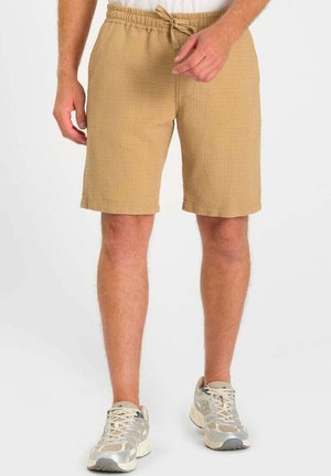 Homme portant un short beige texturé à cordon et des baskets beiges et grises, debout sur un fond blanc.