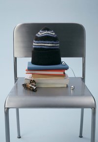 Black and gray striped knit beanie on stacked colorful books, with a toy and small balls on a silver metal chair against a light blue background.