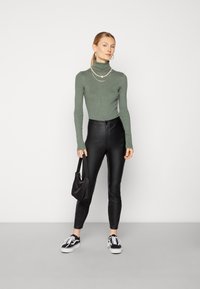 Pullover de gola alta verde com textura canelada, calças pretas de couro sintético de cintura alta, mala preta e ténis pretos. Modelo em pé com as mãos nos quadris.