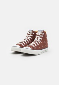 Wysokie trampki Converse w kolorze rdzy z płótna, białe sznurowadła, gumowa podeszwa i wzory w gwiazdy. Posiadają logo na języku i jasnosłoneczne wnętrze.