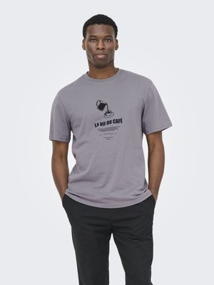 Bărbat purtând un tricou gri cu o cafetieră turnând în ceașcă și textul „La Vie du Café”, asortat cu pantaloni negri, stând cu mâinile în buzunare.