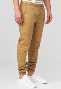 Pantalones jogger marrón claro con cintura y puños elásticos, bolsillos laterales, detalle de cordón, combinados con zapatillas blancas.