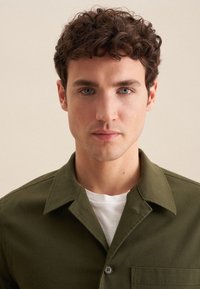 Grüne Baumwollbluse mit einer geknöpften Tasche, ausgestattet mit einem Kentkragen und einer glatten Textur. Darüber getragen wird ein weißes Rundhals-T-Shirt.