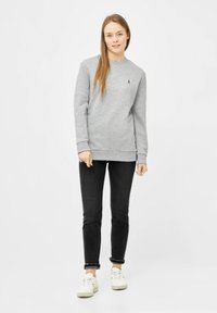 Grauer Sweatshirt mit langen Ärmeln, rundem Halsausschnitt, strukturiertem Stoff und kleinem schwarzen Logo. Kombiniert mit dunklen Jeans und weißen Sneakers.