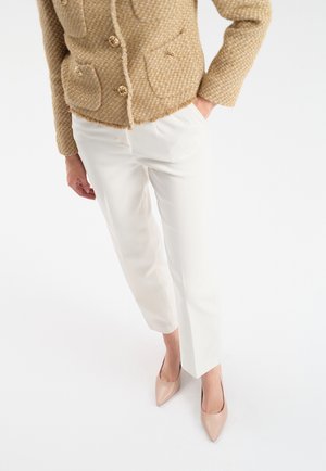 Beżowa, teksturowana kurtka z złotymi guzikami, w połączeniu z białymi spodniami o prostym kroju i cielistymi, szpiczastymi szpilkami, na białym tle.