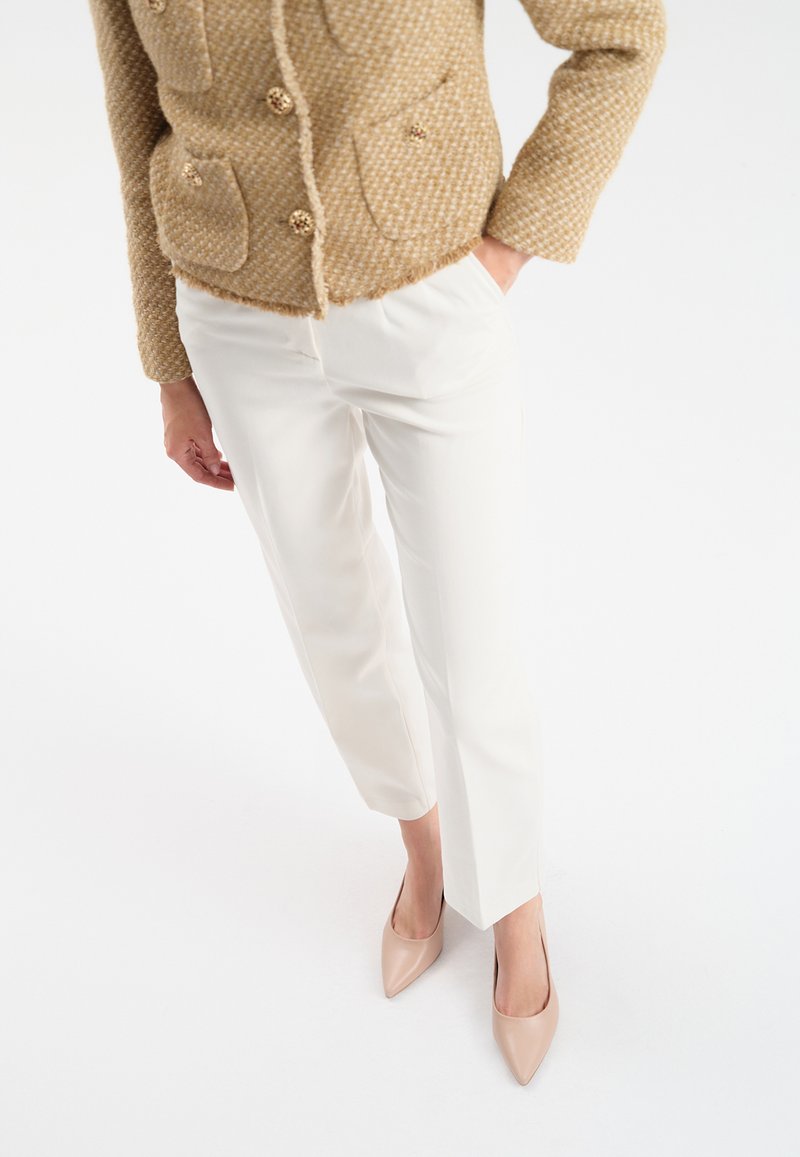 Beżowa, teksturowana kurtka z złotymi guzikami, w połączeniu z białymi spodniami o prostym kroju i cielistymi, szpiczastymi szpilkami, na białym tle.