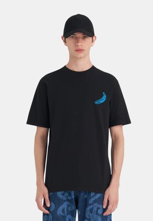 Jeune personne portant une casquette noire et un t-shirt noir avec un motif banane bleu sur la poitrine, associé à un pantalon bleu à motifs.