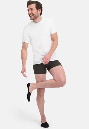 Witte T-shirt met ronde hals, donkergroene boxershorts en zwarte onzichtbare sokken. Model staat met één opgetrokken been, glimlachend, tegen een effen achtergrond.