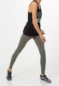 Schwarzes Tanktop mit großem weißem Text, graue Leggings mit strukturierter Optik und schwarze Sneakers mit grauen Akzenten. Modell in Aktionspose.