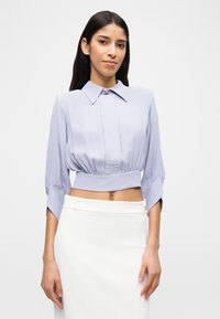 Γυναίκα φορώντας ένα λιλά κοντό μπλουζάκι με φαρδιά γιακά και λεπτομέρεια "EF", συνδυασμένο με μια ψηλόμεση λευκή φούστα, μπροστά σε απλό φόντο.