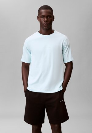 Jonge man met een donkere huid die een lichtblauw shirt met korte mouwen en zwarte korte broek draagt, staand met zijn handen in zijn zakken tegen een effen achtergrond.