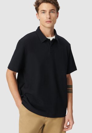 Zwarte poloshirt met een klassieke kraag, korte mouwen en een losse fitting, gecombineerd met khaki-broeken. Glad materiaal en minimale ontwerpdetails.