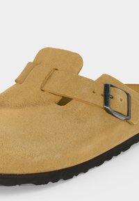 Ανοιχτό καφέ καστόρινο παπούτσι slip-on με ρυθμιζόμενο λουράκι και ασημένιο αγκράφα, με μαύρη υφασμάτινη σόλα και στρογγυλεμένο σχέδιο μύτης.