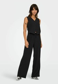 Top noir sans manches et court avec des boutons à l'avant, associé à un pantalon large noir et des bottines argentées métalliques. Tissu lisse, design simple.