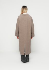 Palton lung, de culoare maro, fabricat dintr-un amestec de lână. Include un guler, mâneci late și o fanta la spate. Purta peste pantaloni gri. Design minimalist.