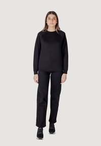 Felpa nera con scollatura a girocollo e maniche raglan, abbinata a pantaloni dritti neri e scarpe da ginnastica nere, in piedi su uno sfondo semplice.