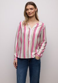 Femme portant un chemisier à manches longues à rayures verticales rose, blanc, beige et bleu, associé à un jean bleu, debout avec une main dans la poche.