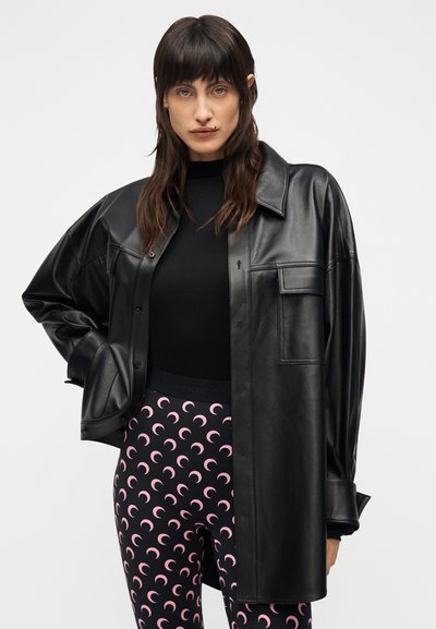 Μαύρο δερμάτινο υπερμεγέθες πουκάμισο-σακάκι με μυτερό γιακά και μπροστινή τσέπη, φορεμένο πάνω από μαύρο τοπ και ροζ κολάν με μοτίβο ημισελήνου.