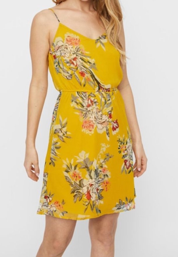 Robe jaune sans manches avec des bretelles spaghetti et un motif floral, portée par une personne debout aux cheveux longs.
