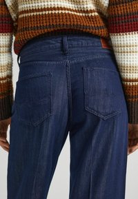 Tmavě modré džíny z denimu se zadními kapsami, s jemnou texturou tkaní a kontrastními švy. Pas je hladký bez viditelných zapínání.