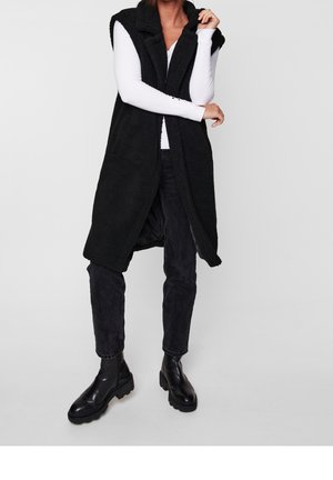 Personne portant une longue veste polaire noire à capuche, une chemise blanche à manches longues, un pantalon noir et des bottines noires sur un fond uni.