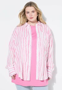 Übergrößiges Button-up-Hemd in Weiß mit rosa wellenförmigen Streifen, über einem einfarbigen rosa T-Shirt getragen. Weicher Stoff, lockere Passform und lange Ärmel.