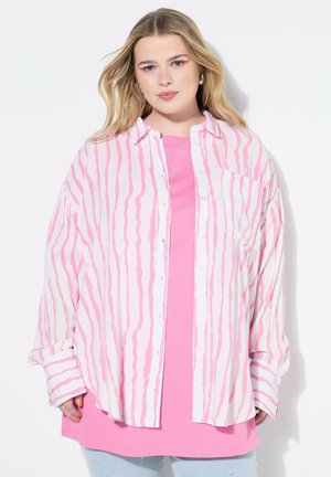 Camicia oversized bianca con strisce ondulate rosa, indossata sopra una maglietta rosa a tinta unita. Tessuto morbido, vestibilità comoda e maniche lunghe.