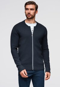Dunkelblauer Reißverschluss-Pullover mit gerippten Bündchen und Saum, ausgestattet mit einem runden Ausschnitt und einer glatten Textur. Getragen über einem weißen Grafik-T-Shirt.