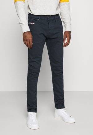Uomo che indossa pantaloni slim-fit blu scuro, sneakers alte bianche e un maglione grigio chiaro con dettagli gialli sulle maniche.