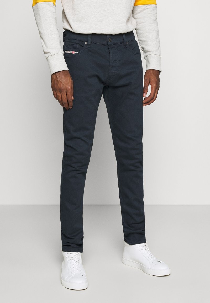 Hombre con pantalones oscuros de corte ajustado, zapatillas altas blancas y un suéter gris claro con detalles amarillos en las mangas.