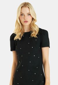 Vestido negro con mangas cortas, tejido texturizado y embellishments de cristal dispersos por el frente, creando un patrón decorativo.
