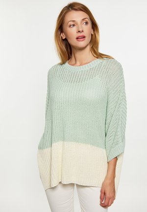 Kvinde iført løsstrikket farveblok-sweater i lysegrøn og creme, der ser let til siden med et neutralt udtryk.