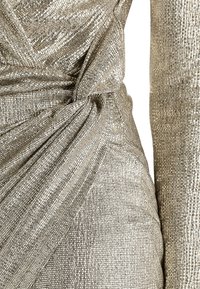 Detail z metalického zlatého šatu s texturovaným, překládaným designem a nařasenou tkaninou, který zdůrazňuje lesk a složitost materiálu.