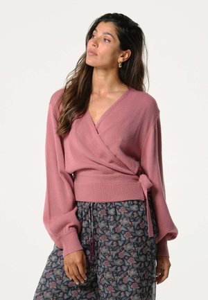 Roze wikkeltrui met een diepe V-hals, wijde mouwen en strikdetail in de taille, gecombineerd met volgepatronen donker bloemenbroek.