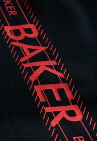 Mustast kangast koosneval taustal on julge punase tekstiga kiri "BAKER" ning seda ümbritseb punaste nõelapistega jooneline muster.