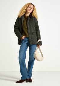 Giovane donna con capelli ricci, indossa una giacca scura e un maglione verde, tiene una borsa crema e sorride in modo rilassato su uno sfondo chiaro.