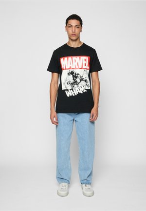 Jeune homme debout portant un t-shirt noir Marvel avec un motif graphique, un jean bleu clair et des baskets blanches sur un fond uni.