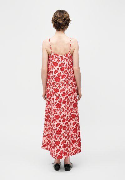 Άτομο με κοντά μαλλιά που φοράει ένα άσπρο αμάνικο φόρεμα μέχρι τον αστράγαλο με κόκκινο floral σχέδιο και μαύρα επίπεδα παπούτσια, στέκεται με την πλάτη προς τα εμάς.