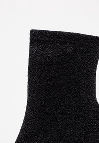 Bota de calcetín negra con un acabado texturizado y brillante. El diseño cuenta con una altura de tobillo ajustada y un borde superior liso.