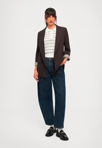 Nainen seisoo kädet taskuissa ruskeassa bleiserissä, raidallisessa lyhythihaisessa polo paidassa, leveälahkeisissa farkuissa, valkoisissa sukissa ja mustissa loafer-kengissä.